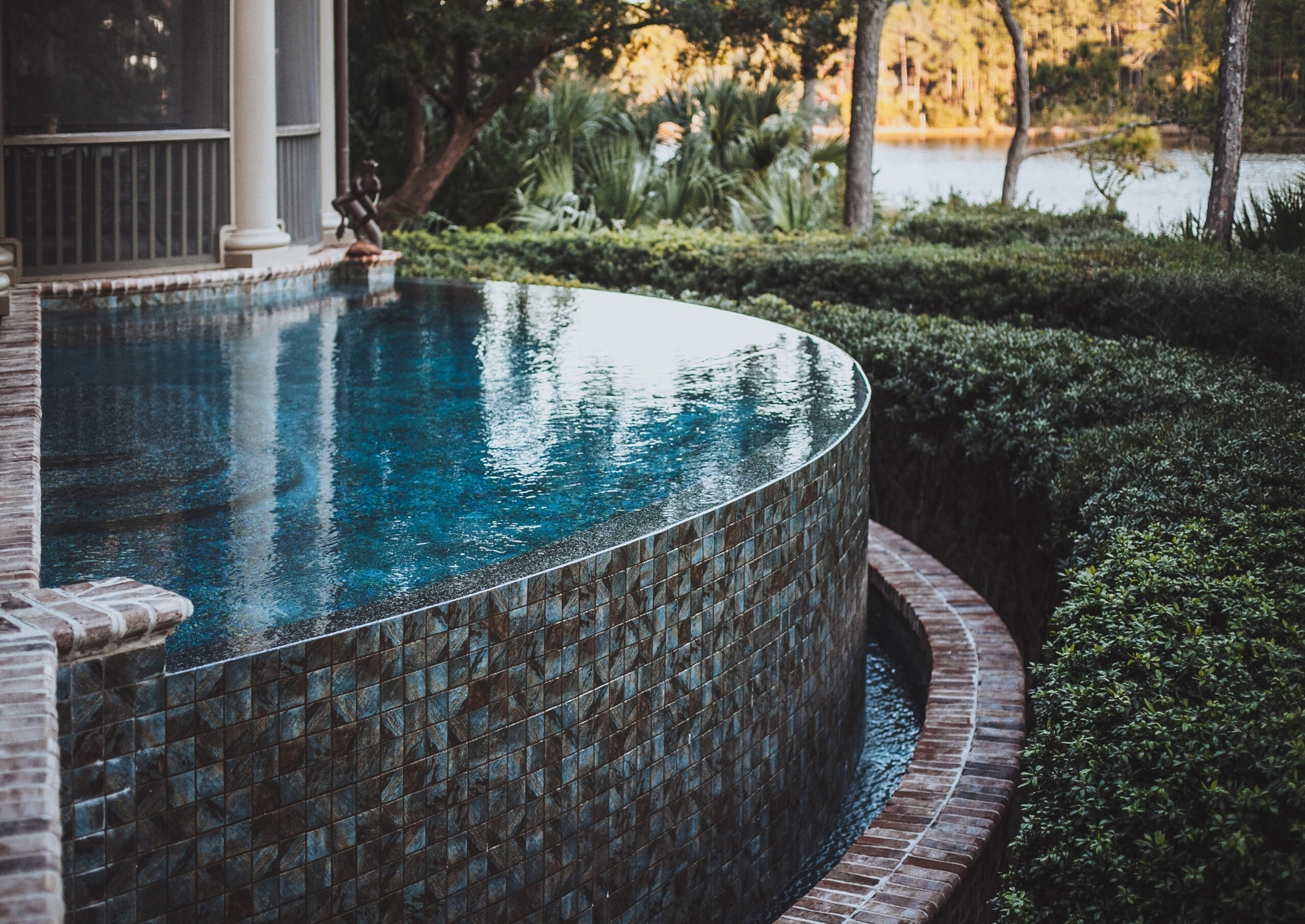 Luxurious infinity-edge pool with stone-tiled exterior, set beside a covered patio with columns, overlooking lush greenery and a serene water view in the background.