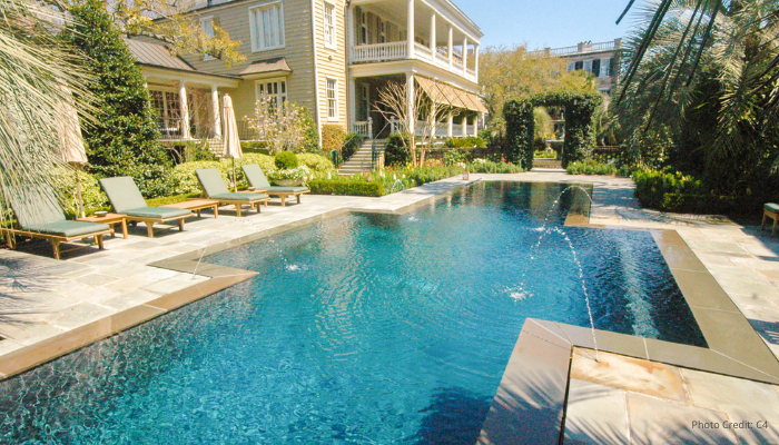 Elegant backyard pool with integrated spa, surrounded by lounge chairs and lush greenery, set beside a large two-story home with a covered patio and landscaped garden.