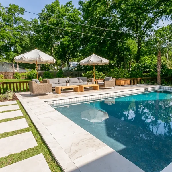 Charming backyard pool with neutral patio furniture, umbrellas, and lush landscaping.