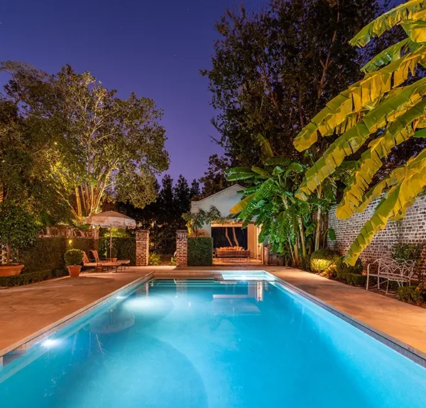 Tranquil backyard pool at night with ambient lighting and tropical foliage.