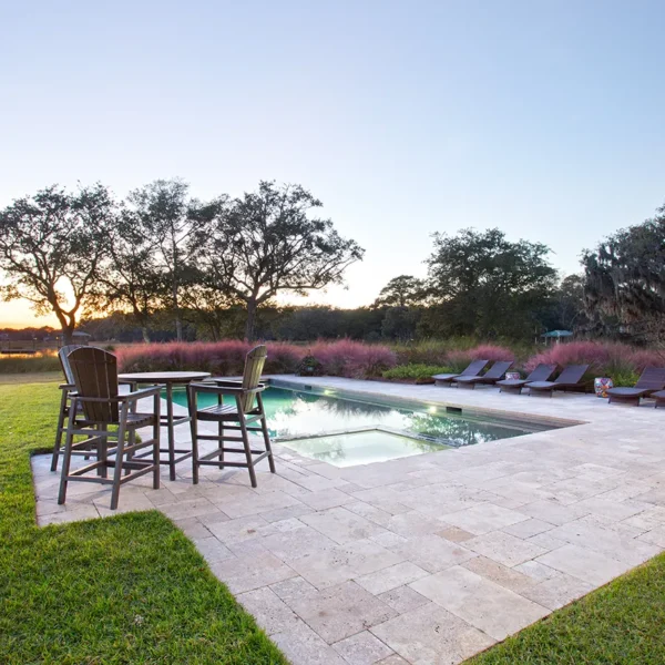 Backyard swimming pool with lounge chairs, dining table, and waterfront view at sunset.