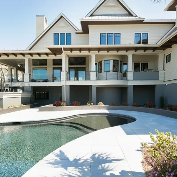 Back view of a coastal-style luxury home with a curved-edge pool and clean white stone decking, framed by tropical landscaping.