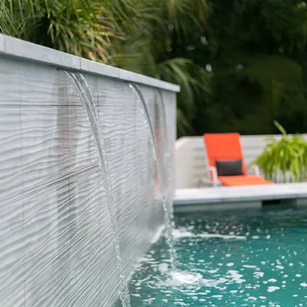 Close-up of a pool waterfall feature with lounge chairs in the background.